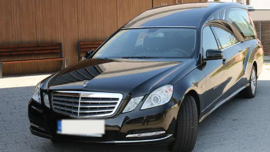 Czarny karawan zaparkowany przed budynkiem domu pogrzebowego, przy drewnianej elewacji.